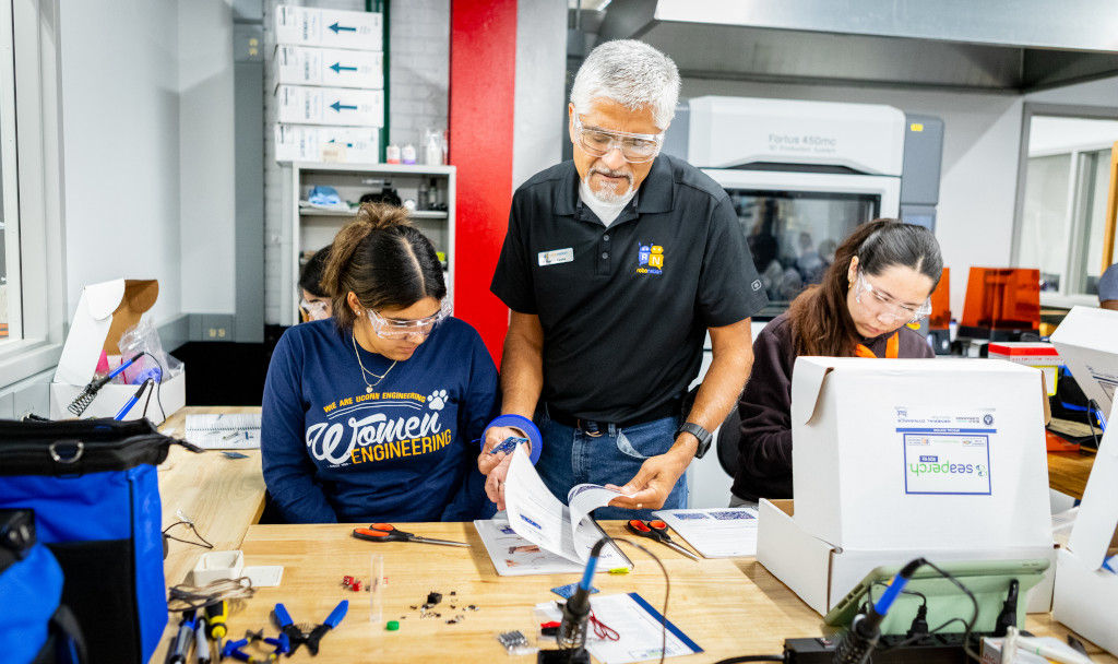 A male teacher showing two female students a manual on how to conduct a certain operation with electronic tools.